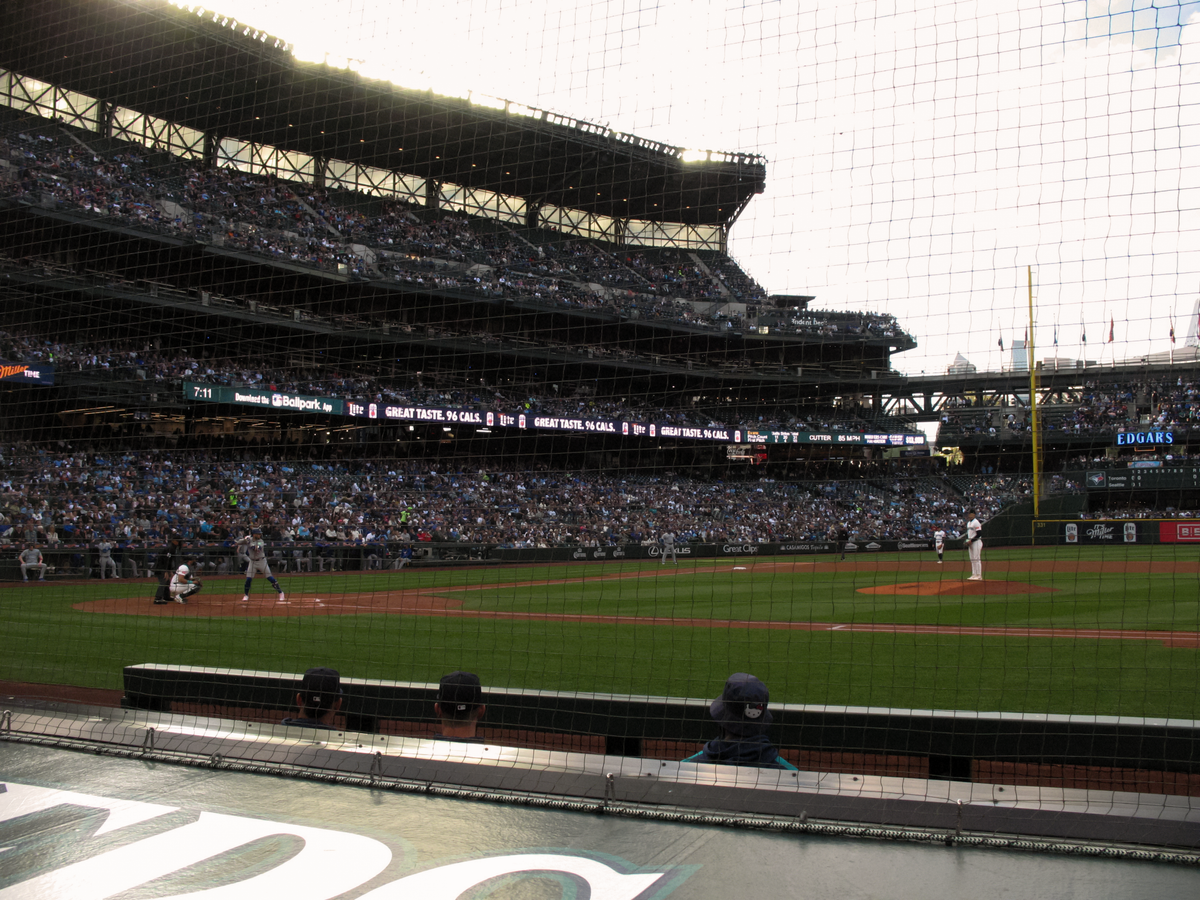 A side view of home plate and the pitcher's mound at T-Mobile Park, with Bo Bichette ready to bat and Logan Evans on the mound. The background shows the stadium filled with fans.